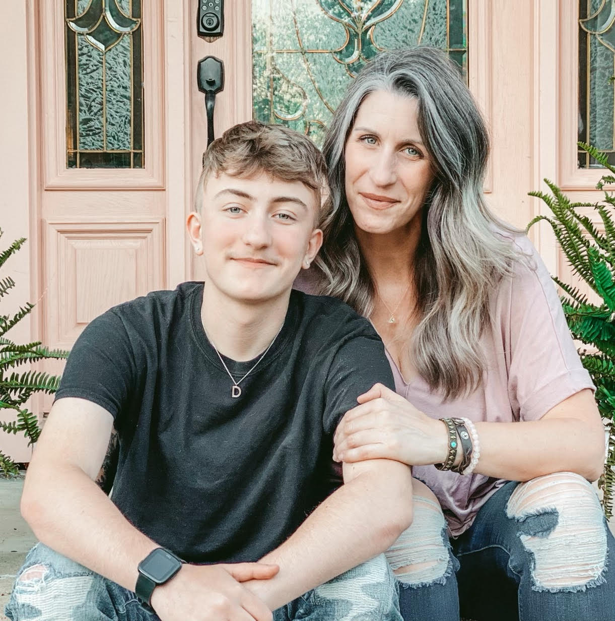 Dylan, a white boy in a black tshirt with blonde hair, and his mom, Joanna, a white women with gray hair and a pink tshirt sitting on a poarch with a peach-painted wood and green stained glass windows.